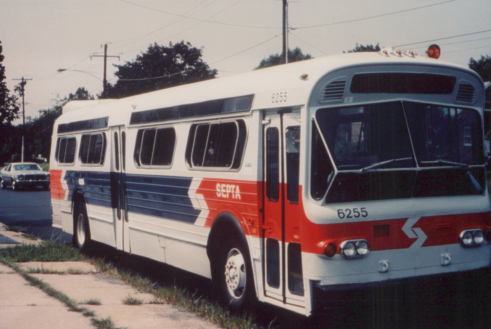 A white, red, and blue SEPTA bus with the number 6255 displayed on the front, side, and roof, parked on a street with a car in the background and trees lining the road.