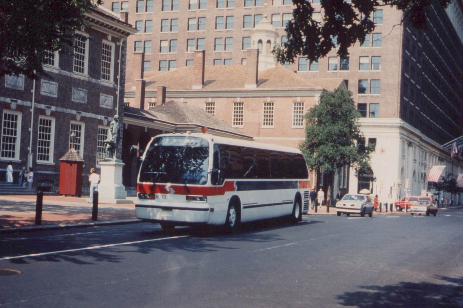 A white and red bus drives on a city street past historic brick buildings on a sunny day, with pedestrians on the sidewalk.