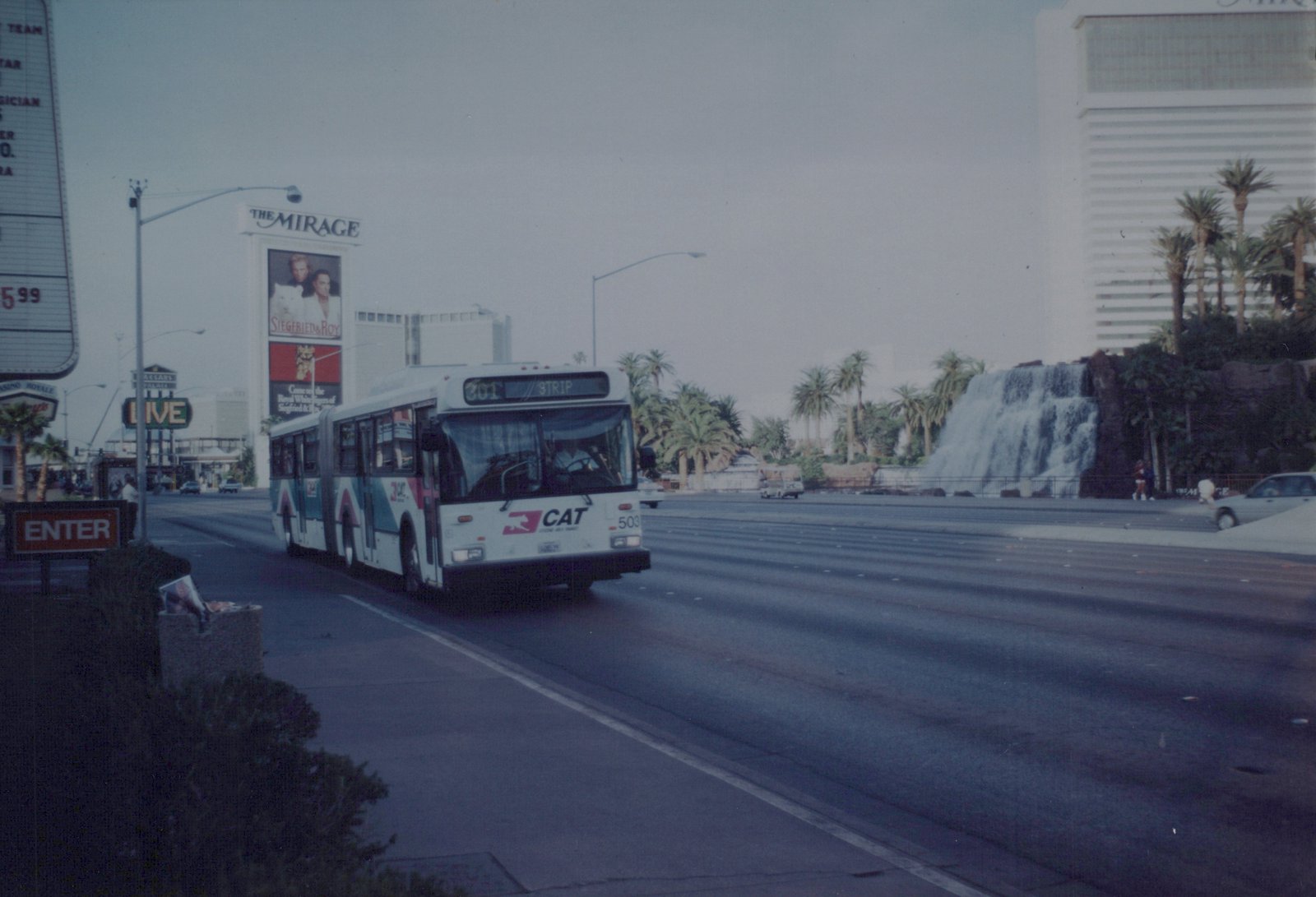 A white and teal CAT bus numbered 503 on a Las Vegas street, with The Mirage hotel and waterfall visible behind it, under an overcast sky.