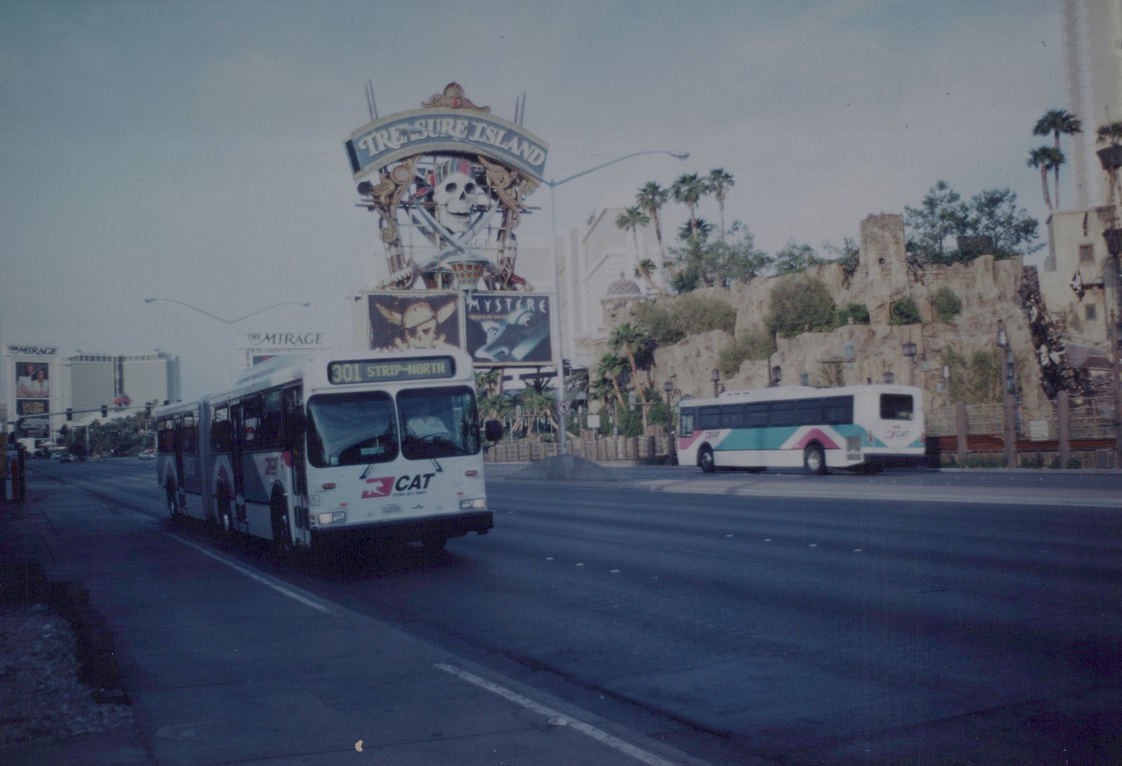 A white articulated bus with pink and teal stripes, marked CAT, drives on a wide city street with a large pirate-themed sign for Treasure Island in the background.