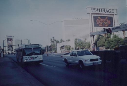 A white CAT bus numbered 618, not in service, is on a city street with The Mirage hotel and a white pickup truck visible. The sky is overcast.