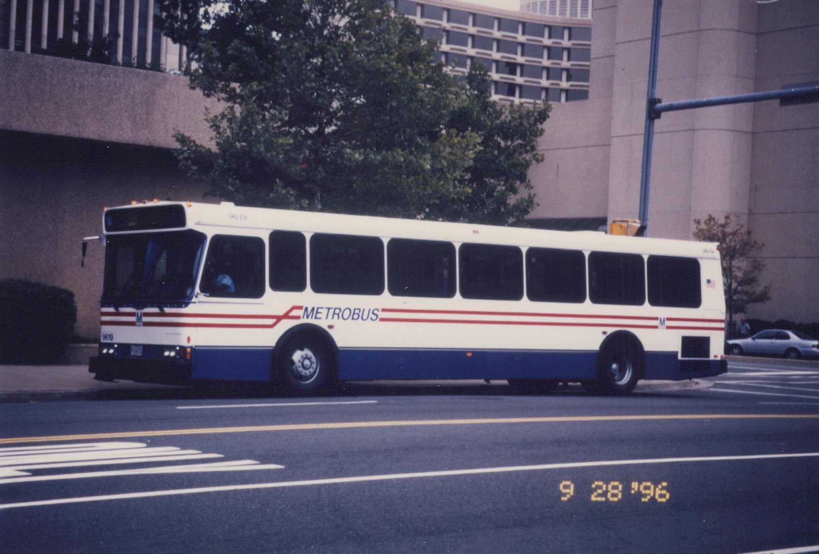 A white and blue Metrobus with red stripes, number 9619, parked on a city street with a building and trees in the background, dated September 28, 1996.