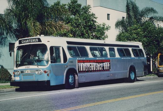 A white and blue Santa Monica Municipal Bus Lines motor bus, number 5180, is parked on a street with palm trees in the background; it features a 1986 Independence Day movie ad on its side.