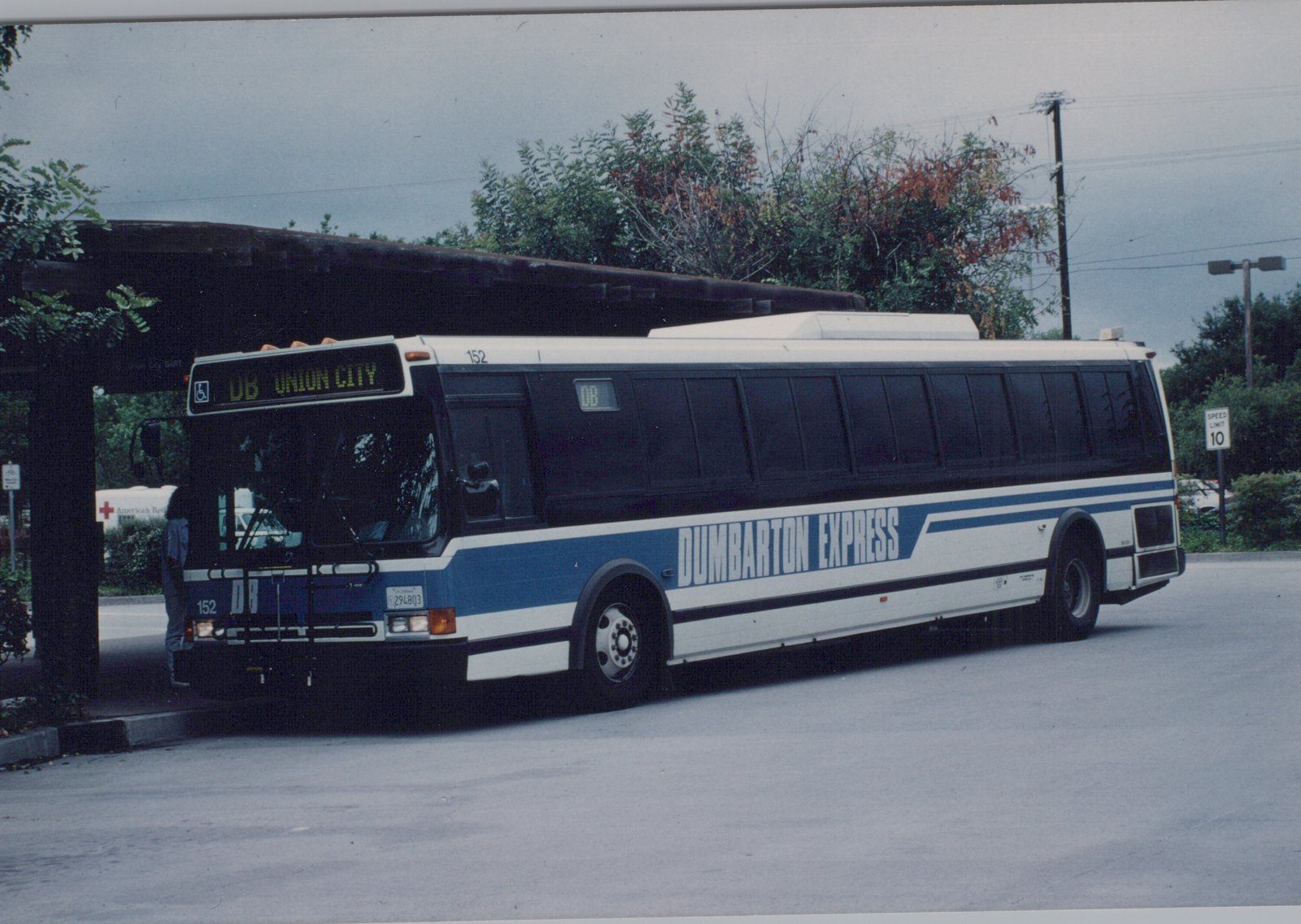 A white and blue Dumbarton Express motor bus, number 152, is parked at a bus stop under an overcast sky with autumn foliage in the background.