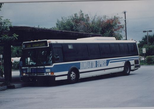 A white and blue Dumbarton Express motor bus, number 152, is parked at a bus stop under an overcast sky with autumn foliage in the background.