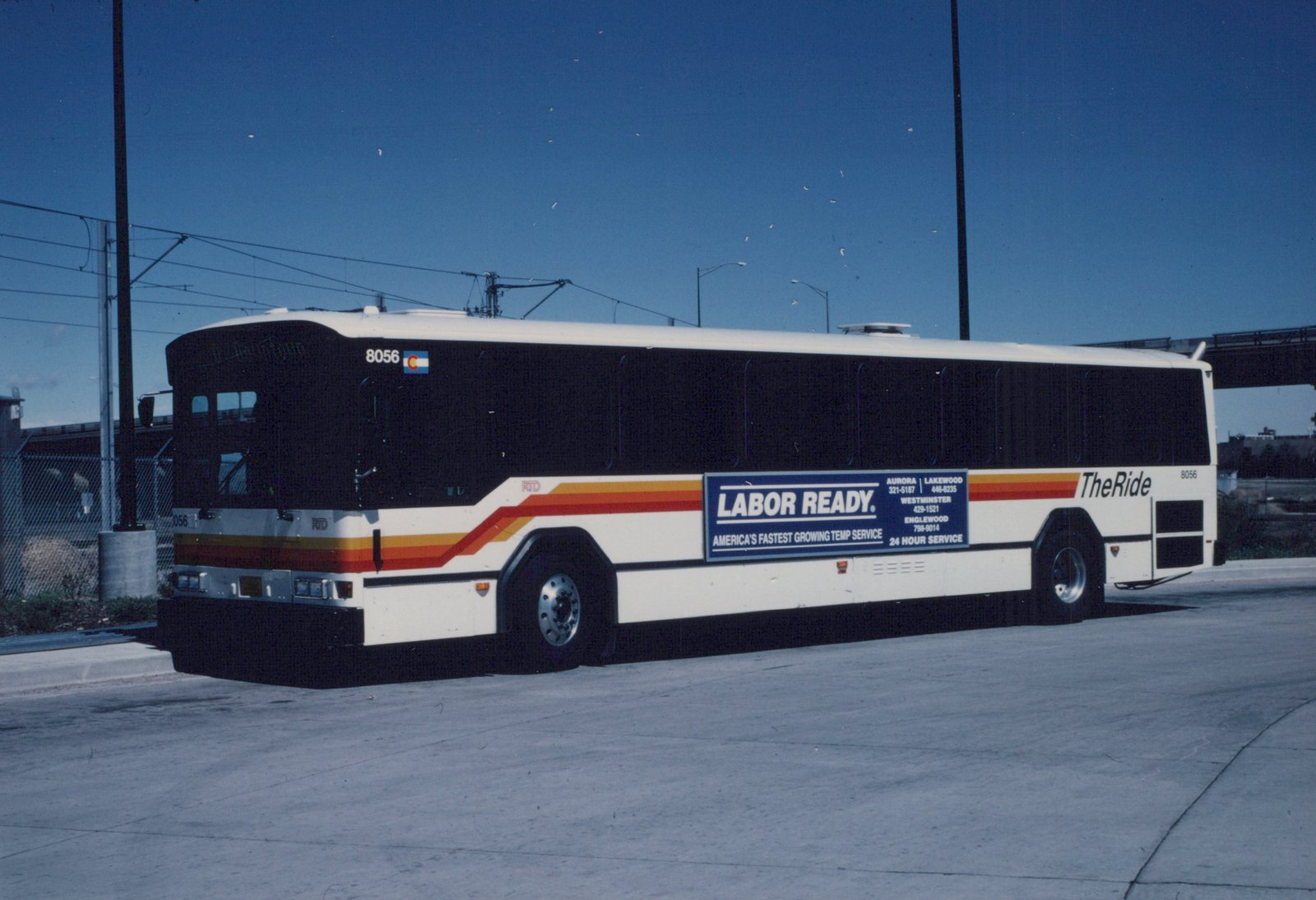 Side view of a white RTD motor bus, number 8056, with a 'LABOR READY' advertisement, parked on a paved lot under a clear blue sky with overhead wires.
