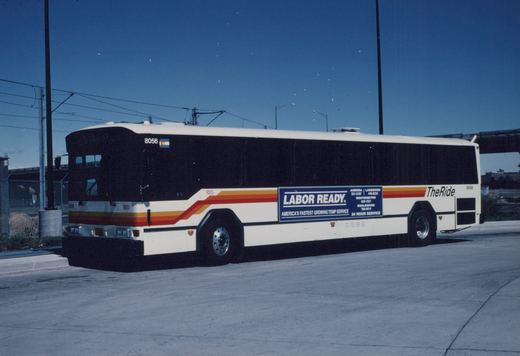 Side view of a white RTD motor bus, number 8056, with a 'LABOR READY' advertisement, parked on a paved lot under a clear blue sky with overhead wires.