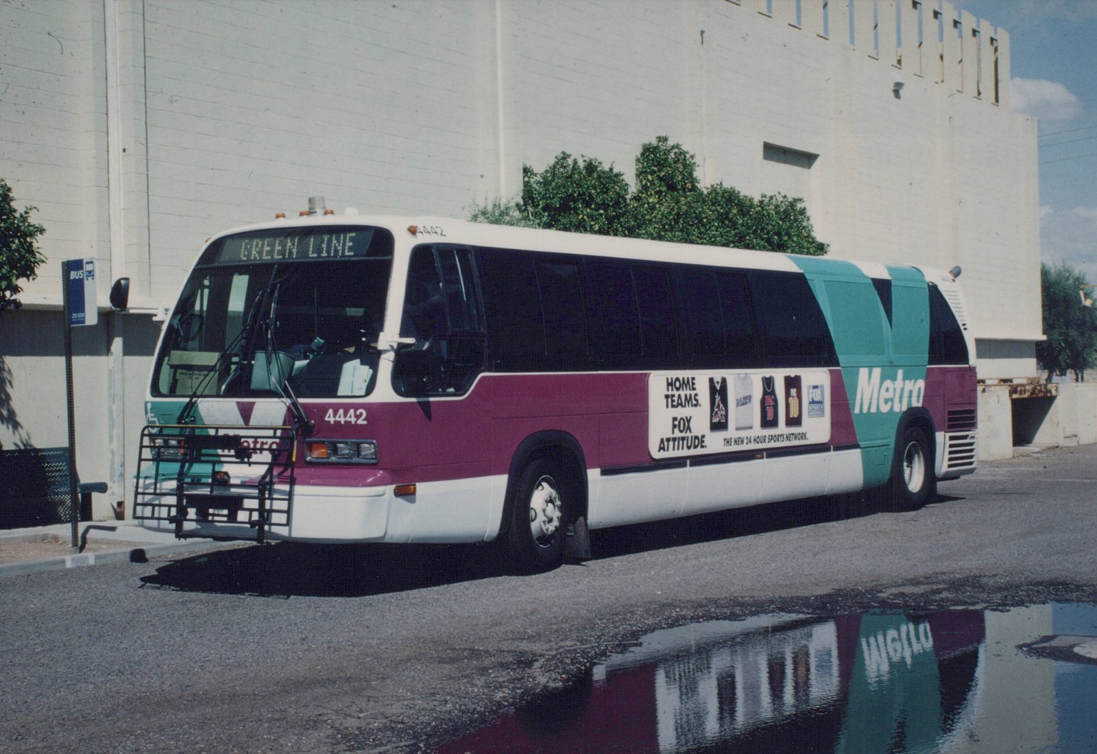 A maroon, teal, and white Valley Metro bus numbered 4442 with a 'Green Line' destination sign is parked at a bus stop next to a puddle reflecting its livery, with a large white building in the background.