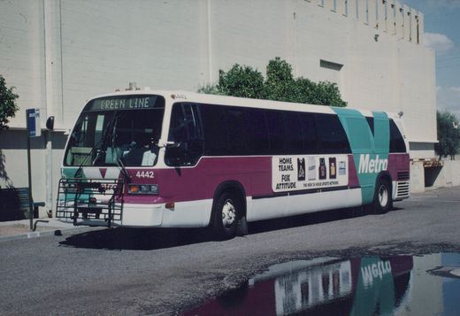 A maroon, teal, and white Valley Metro bus numbered 4442 with a 'Green Line' destination sign is parked at a bus stop next to a puddle reflecting its livery, with a large white building in the background.