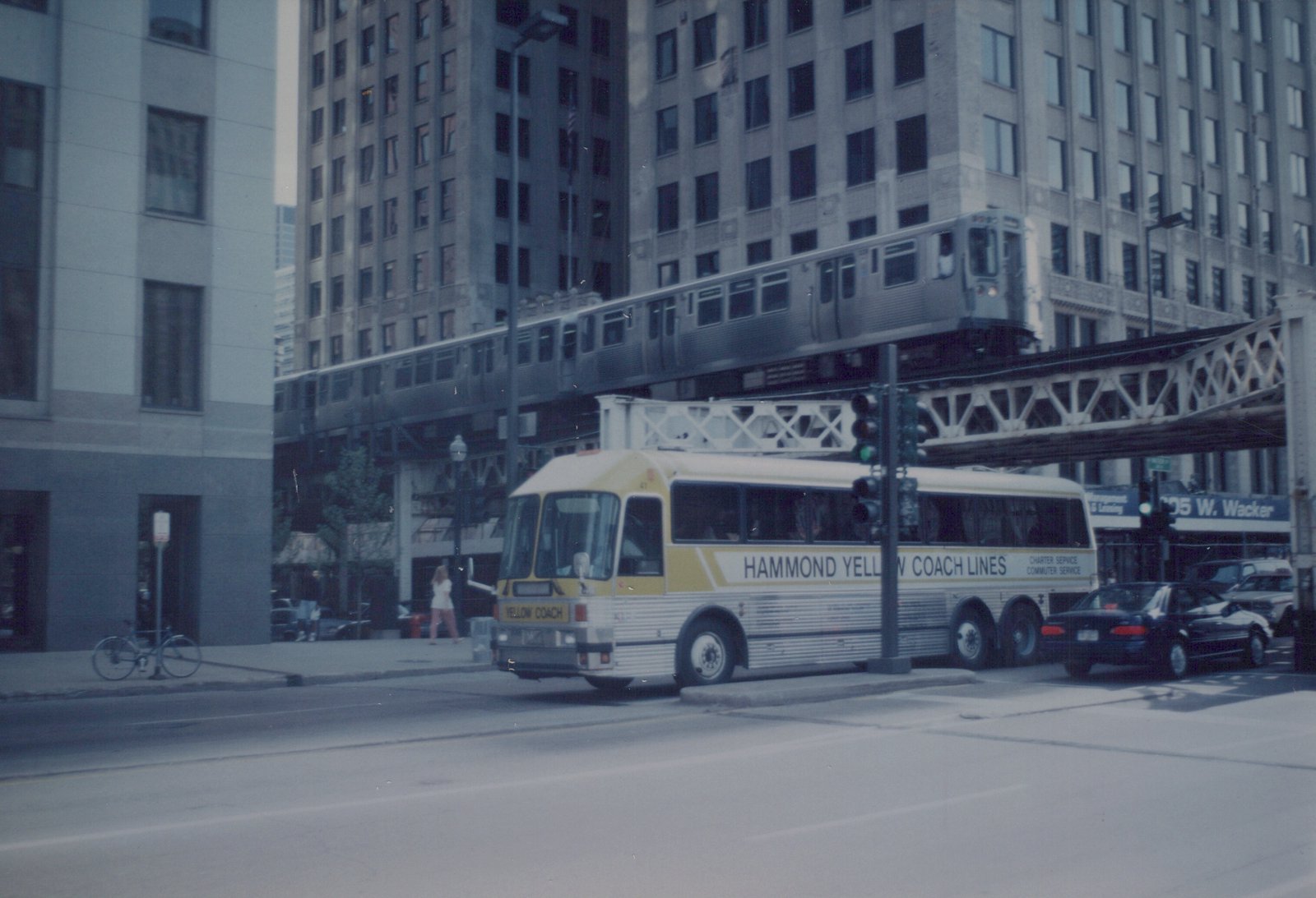 A yellow and white bus with 'Hammond Yellow Coach Lines' on its side is stopped on a city street as a silver rapid transit train travels on an elevated track above it.