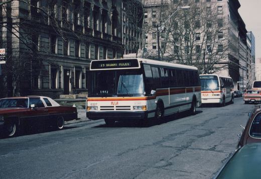 Two Regional Transportation Authority buses, 8327 and 8237, are parked on a city street lined with bare trees and buildings, with vintage cars nearby, on a clear winter day.