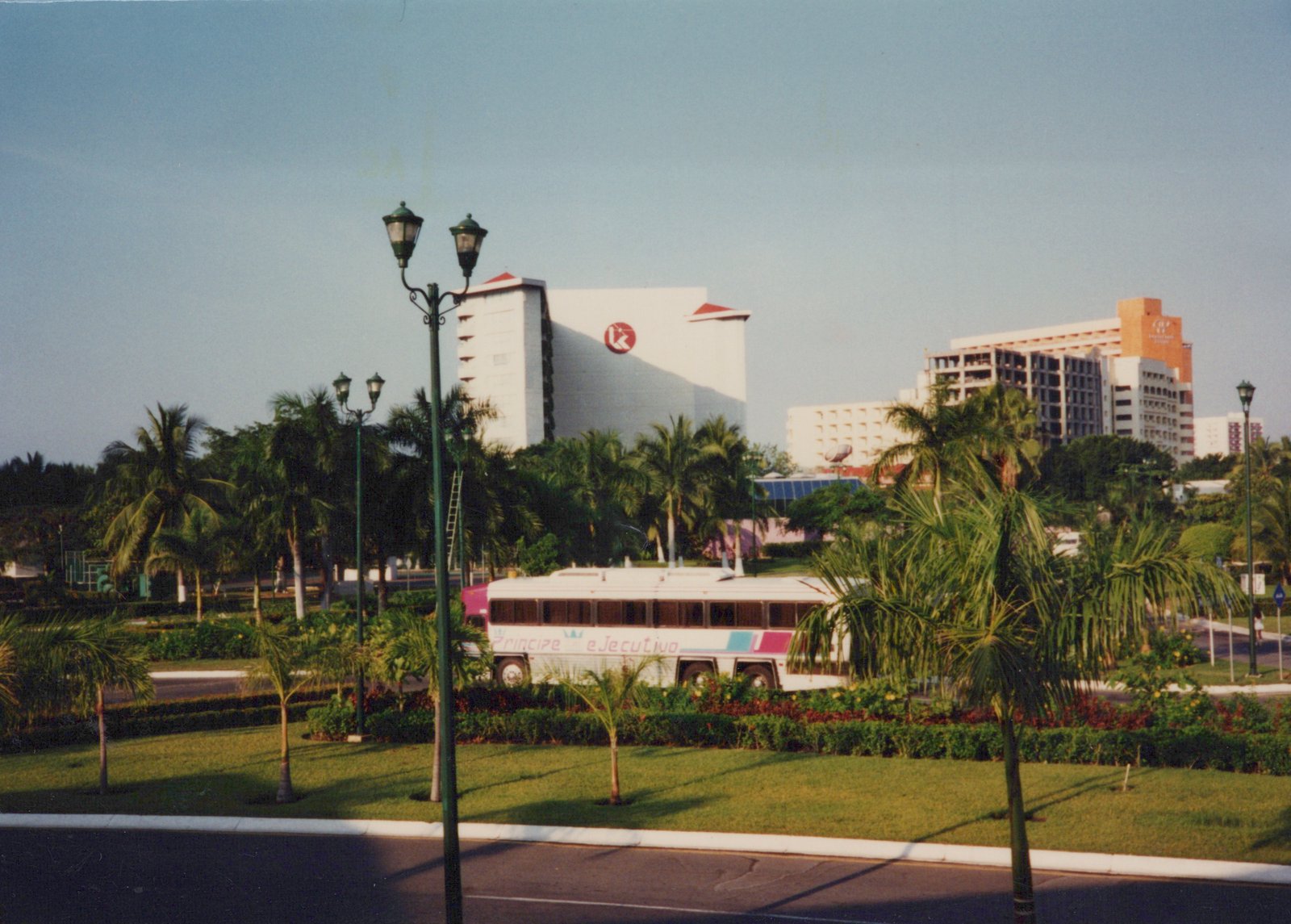 A white bus with 'Principe Ejecutivo' lettering on its side is parked near palm trees and manicured lawns, with large resort hotels in the background under a clear blue sky.