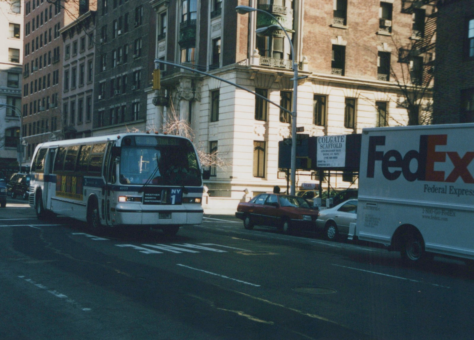 A white and blue MTA bus with the number 4803 and route M5 to Houston Street is driving down a city street with a FedEx truck and other cars nearby.