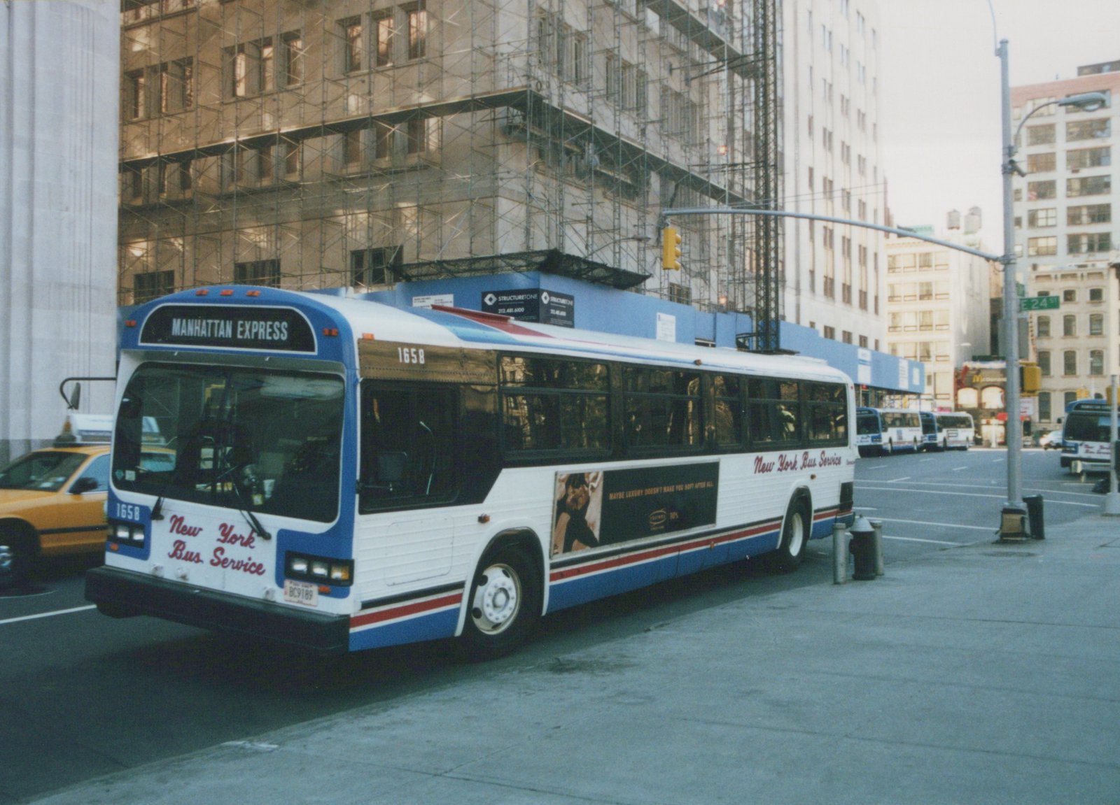 A white, blue, and red New York Bus Service motor bus, number 1658, labeled 'Manhattan Express,' is parked on a city street with scaffolding-covered buildings in the background.