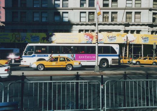 A white and blue MTA bus with the number 4151 on its side is parked on a city street, with a yellow taxi cab in front and a large movie banner for 'Rugrats in Paris' on the building behind it.