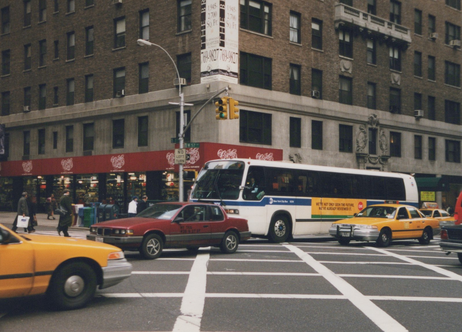 A white and blue New York City Bus with the number 9226 is at a city intersection with yellow taxis and pedestrians, under an overcast sky, in front of a large brick building with a 'Douglas Elliman' sign.
