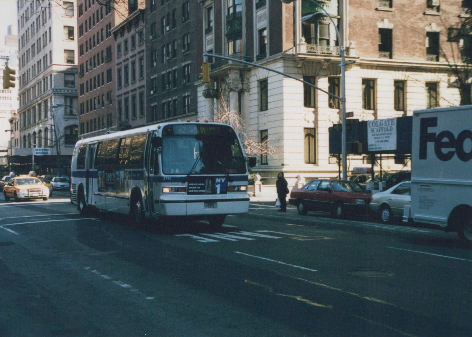 A city bus, marked M2 9th Village, drives down a street lined with tall buildings and parked vehicles on a clear winter day.
