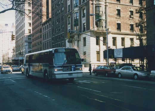 A white and blue bus with the number 8801 and route M2 via 5th Avenue is driving down a city street lined with tall buildings and parked cars.