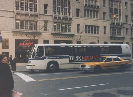 A white and blue MTA bus with the number 9194 is driving down a city street next to a yellow taxi; pedestrians walk on the sidewalk and a store with a neon sign reading 'Pick A Bagel' is visible in the background.