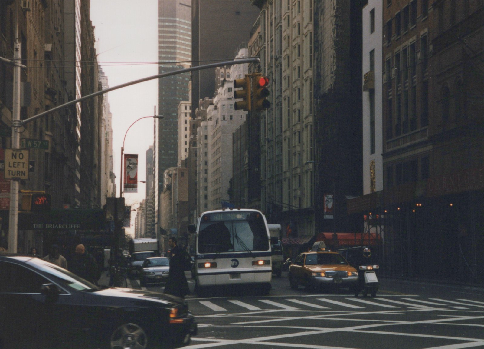 A white and blue MTA bus, number 9216, stopped at an intersection on a cloudy day in New York City, with a yellow taxi and pedestrians nearby.