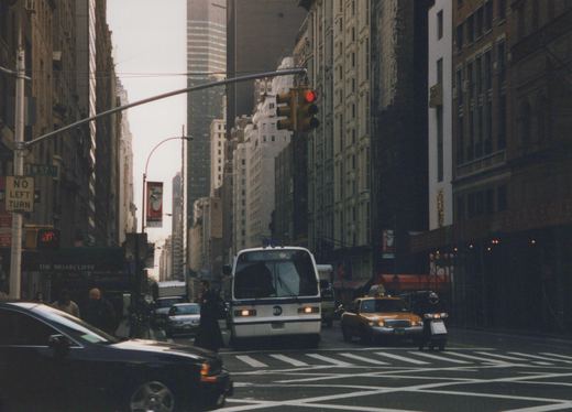 A white and blue MTA bus, number 9216, stopped at an intersection on a cloudy day in New York City, with a yellow taxi and pedestrians nearby.