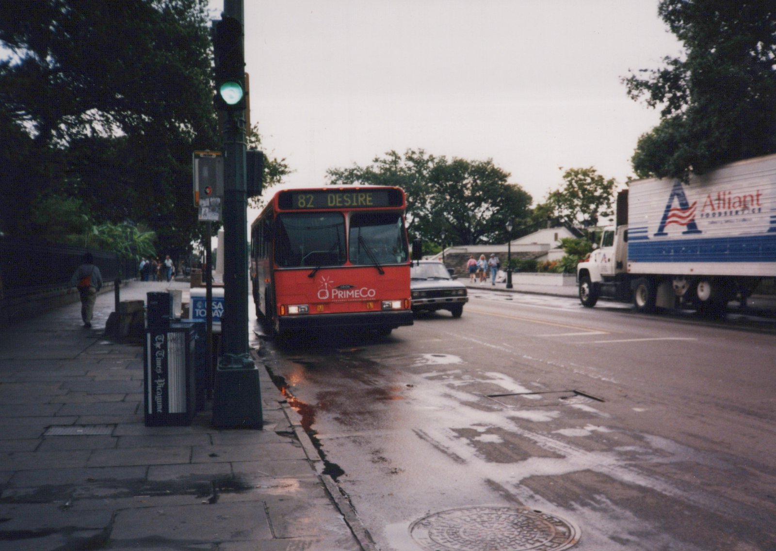 A red city bus with the sign '82 DESIRE' is stopped at a wet street corner under a green traffic light, with a newspaper box for 'The Times-Picayune' on the sidewalk and a white delivery truck nearby.