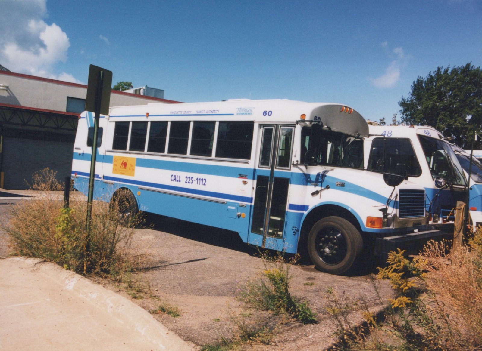 A white and blue motor bus with the number 60 and 'Marquette County Transit Authority' lettering is parked on a gravel lot. Another bus with the number 48 is behind it. The sky is blue and clear.