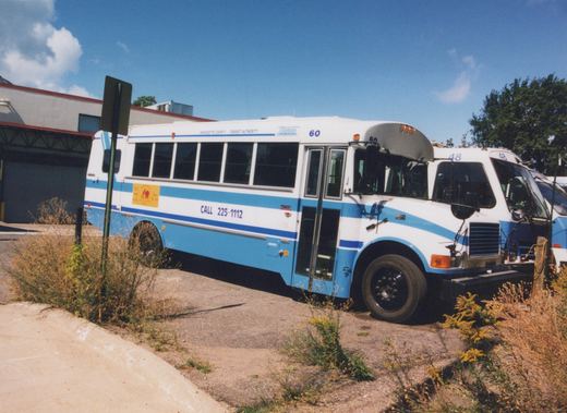 A white and blue motor bus with the number 60 and 'Marquette County Transit Authority' lettering is parked on a gravel lot. Another bus with the number 48 is behind it. The sky is blue and clear.
