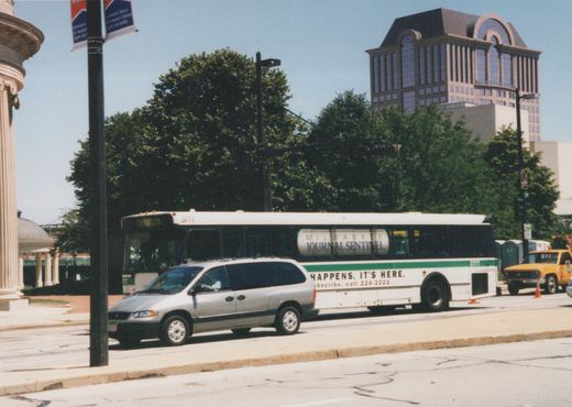A white and green motor bus with the number 3777 is parked on a city street, featuring a Milwaukee Journal Sentinel ad on its side, with a silver minivan in front and a modern office building in the background.