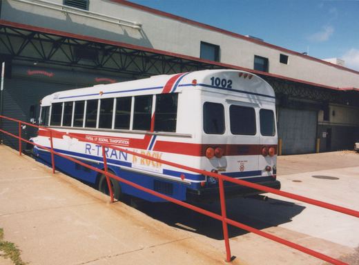 A white, red and blue motor bus numbered 1002, marked for Central U.P. Regional Transportation, is parked outside a garage with red railings under a clear sky.