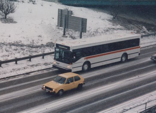 A white Metro Transit bus with a red stripe and the number 1211 is driving on a snowy highway, with a yellow car and another vehicle visible in adjacent lanes.
