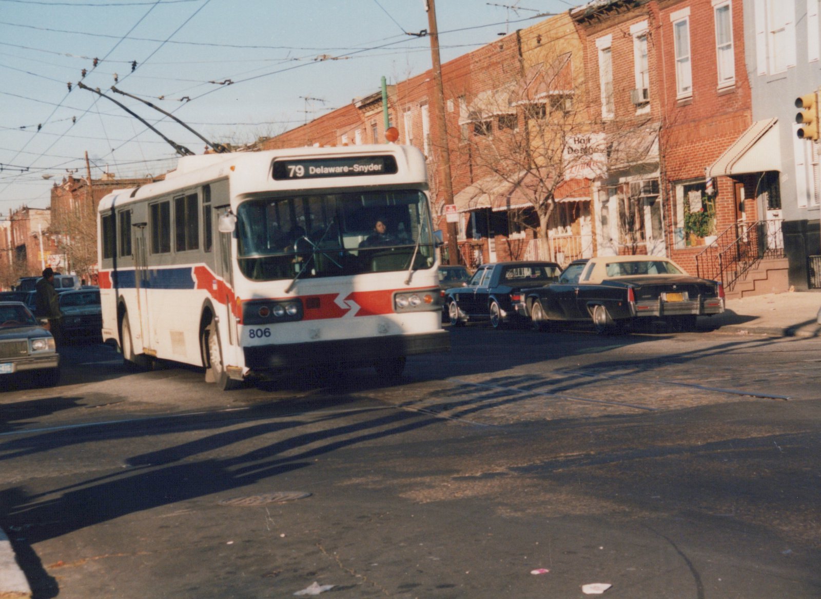 A white, red, and blue trolleybus with the number 806 and destination sign reading '79 Delaware-Snyder' is driving down a city street lined with brick buildings and parked cars.