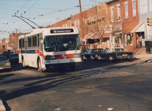A white, red, and blue trolleybus with the number 806 and destination sign reading '79 Delaware-Snyder' is driving down a city street lined with brick buildings and parked cars.
