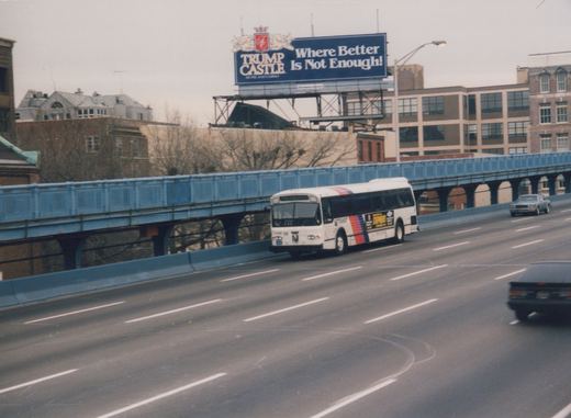 A white NJ Transit bus with the number 7446 is driving on an elevated highway under a large blue billboard that reads 'Trump Castle: Where Better Is Not Enough!', with a blue metal bridge structure overhead and cars in the background.