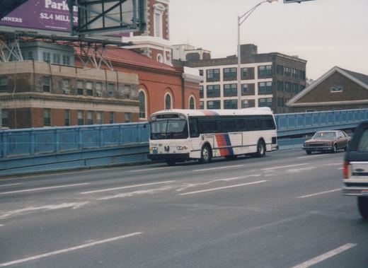 A white NJ Transit bus with a tri-color stripe crosses a blue bridge, with city buildings and other cars in the background.