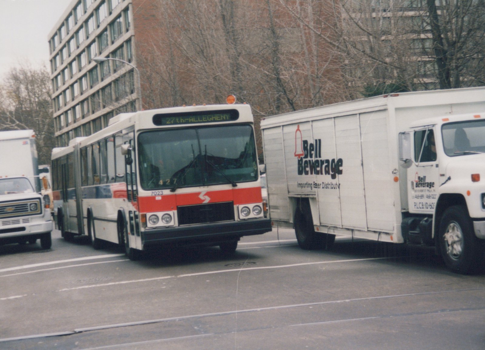 A white and red articulated bus with the number 7023 is driving on a city street, next to a white delivery truck with 'Bell Beverage' lettering, with bare trees and a building in the background.