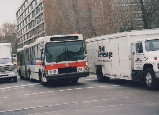 A white and red articulated bus with the number 7023 is driving on a city street, next to a white delivery truck with 'Bell Beverage' lettering, with bare trees and a building in the background.
