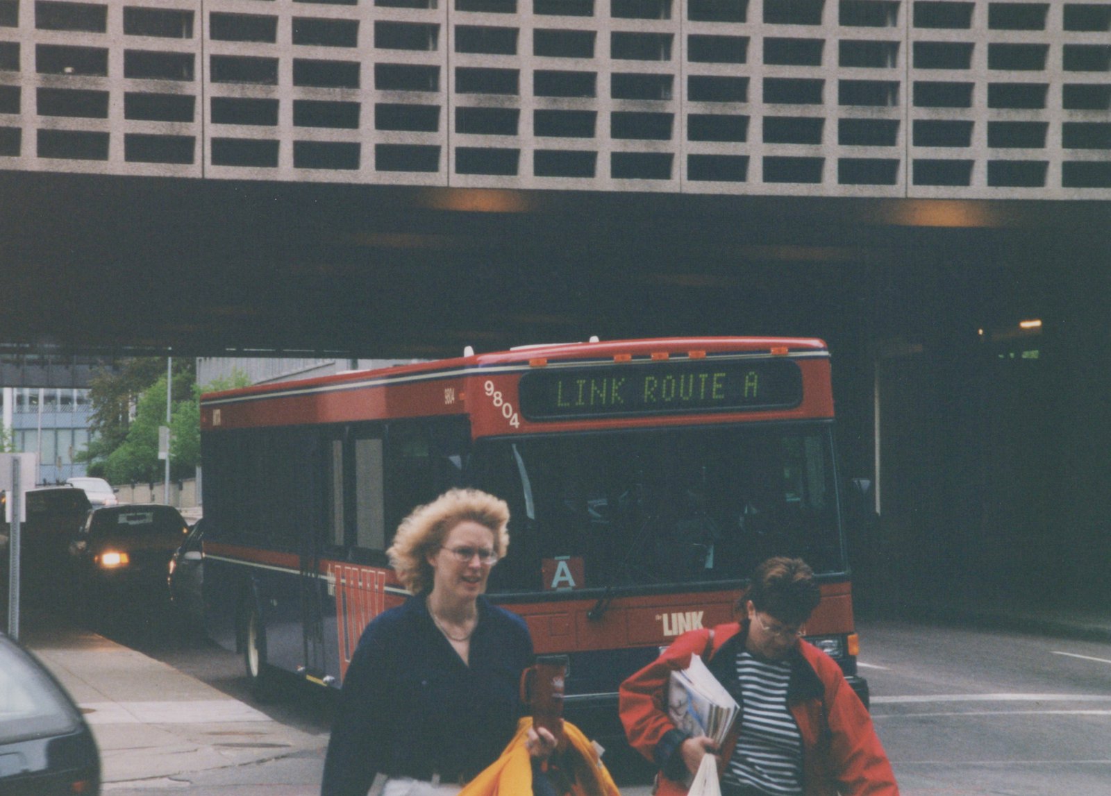 A red and black city bus with 'LINK ROUTE A' on the front sign is driving on a street under a large concrete structure; two women are walking on the sidewalk in the foreground.