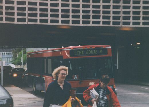 A red and black city bus with 'LINK ROUTE A' on the front sign is driving on a street under a large concrete structure; two women are walking on the sidewalk in the foreground.