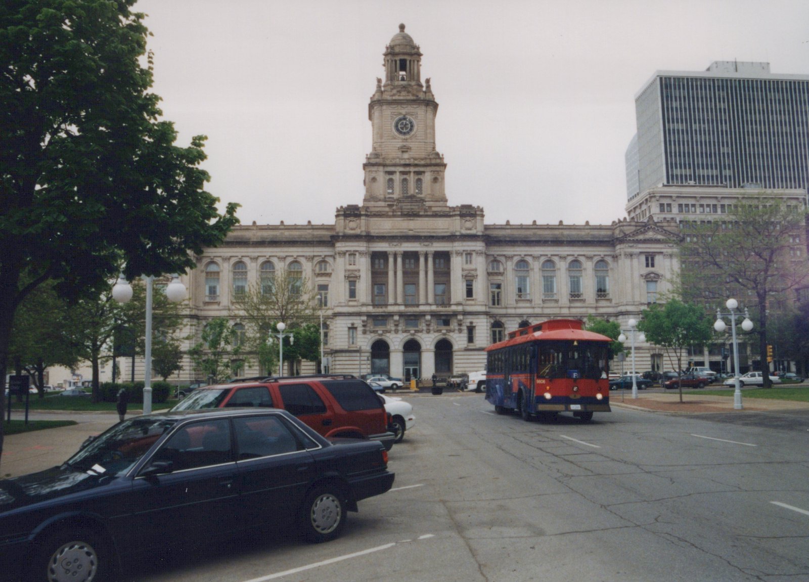 A red and blue trolleybus with the number 9806 drives down a city street, with a large stone building and a clock tower in the background. Several cars are parked nearby.