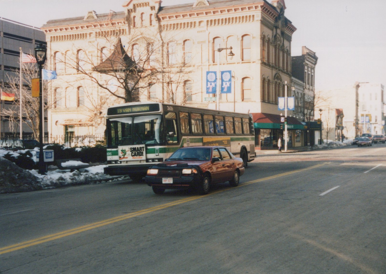 A white and green city bus with the number 3606 and destination '19 Van Norman' is on a city street with a red car in front of it; snow is on the ground and banners reading 'JOY' hang from a building in the background.