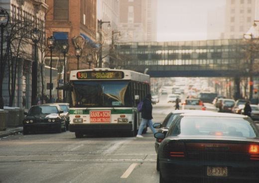 A white and green city bus with the number 4111 on the back, driving on a street with cars and pedestrians, under an overpass.