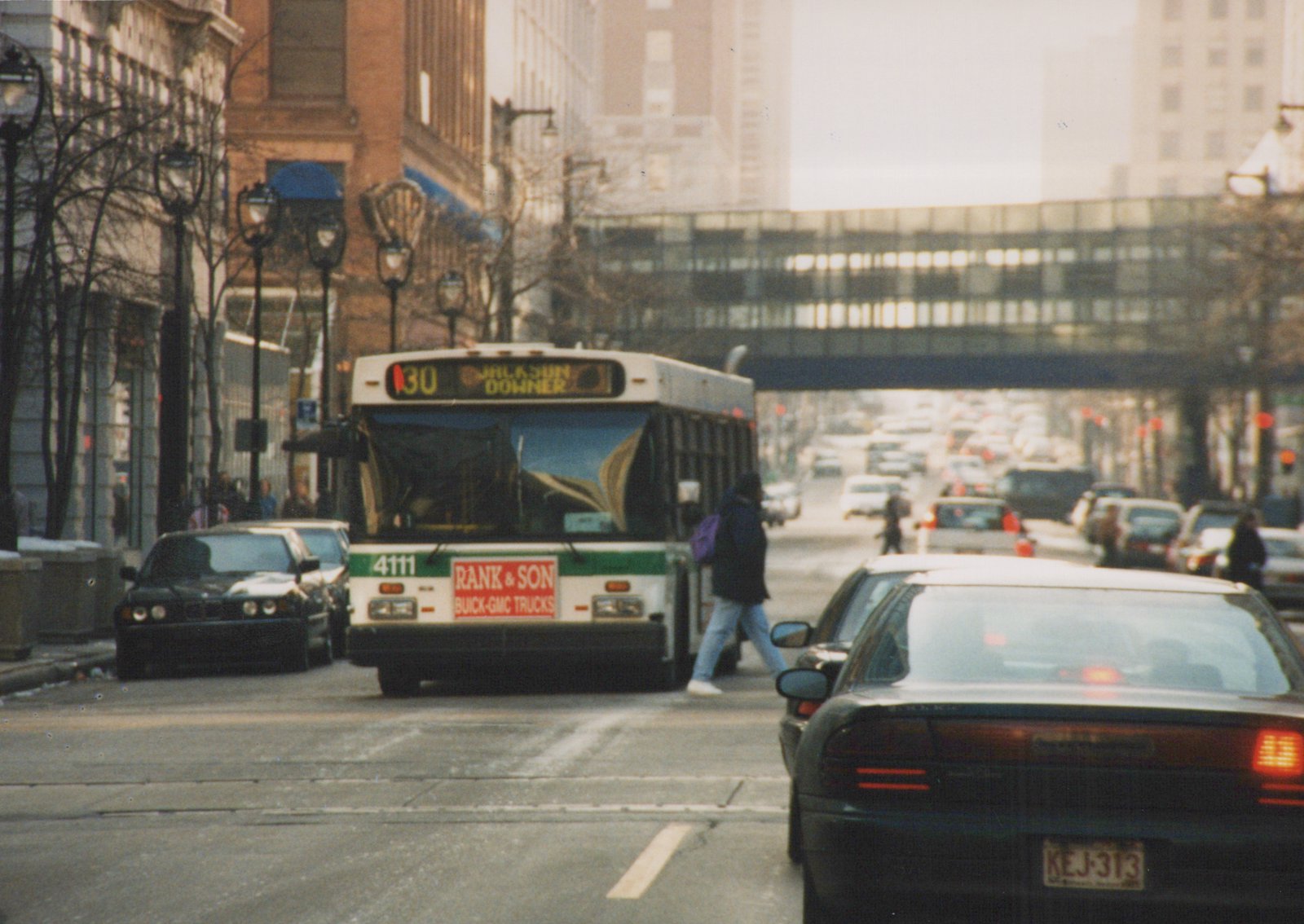 A white and green city bus with the number 4111 on the back, driving on a street with cars and pedestrians, under an overpass.