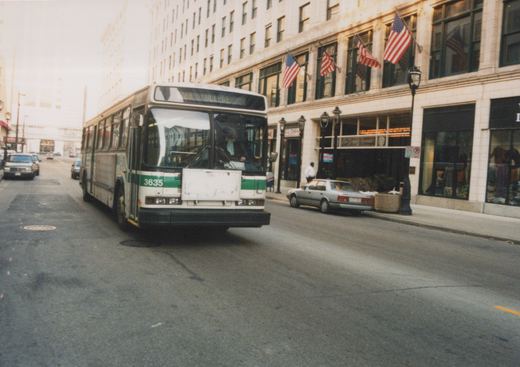 A white and green motor bus with the number 3635 on the front is driving down a city street lined with tall buildings and American flags.