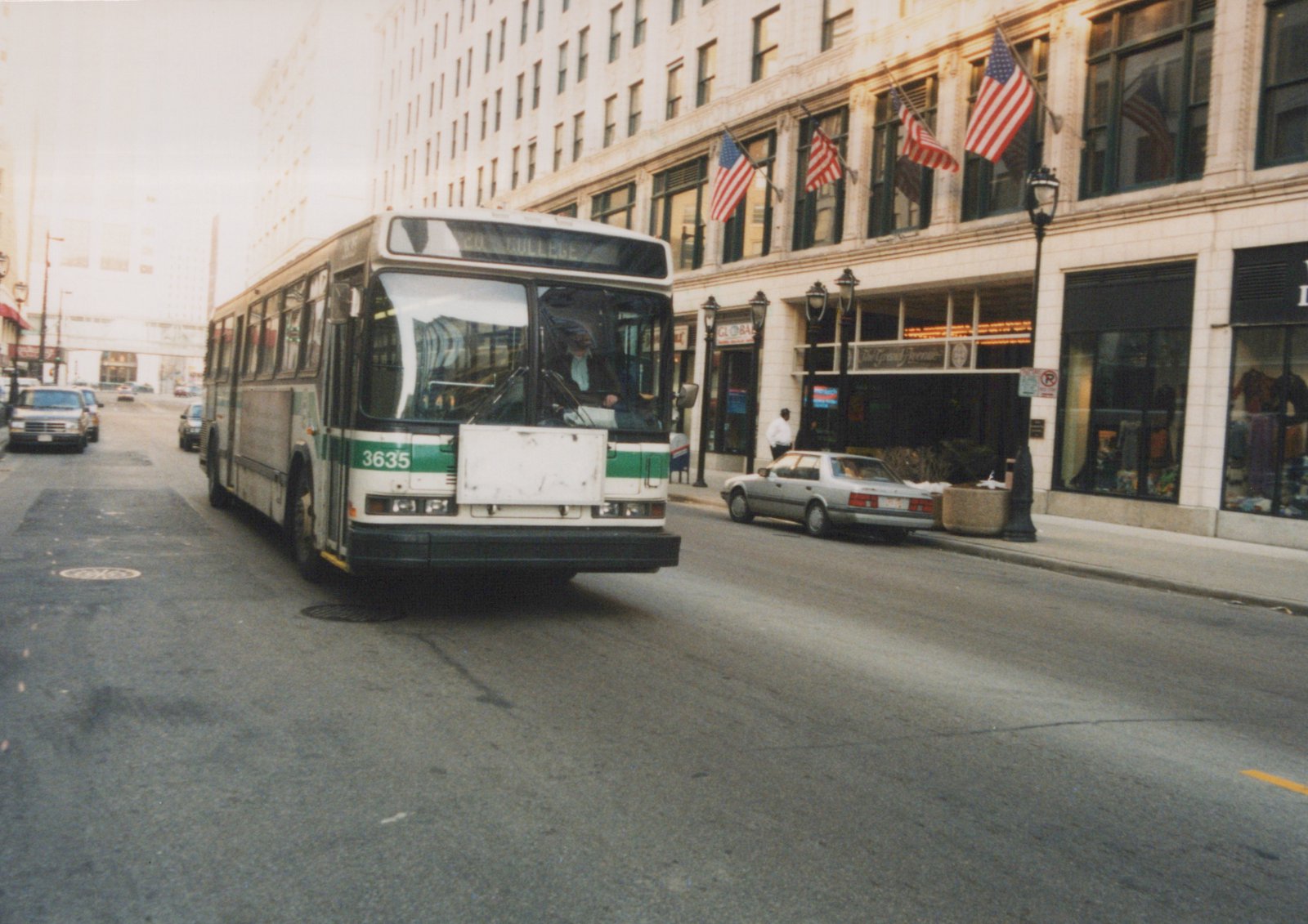 A white and green motor bus with the number 3635 on the front is driving down a city street lined with tall buildings and American flags.