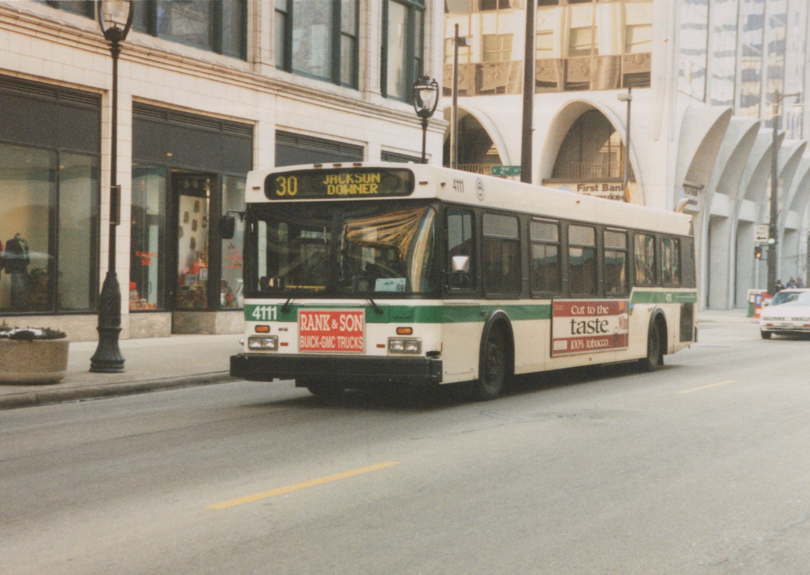 A white and green Flxible Metro bus with the number 4111, operated by Milwaukee County Transit System, is driving on a city street with a '30 Jackson Downer' destination sign.