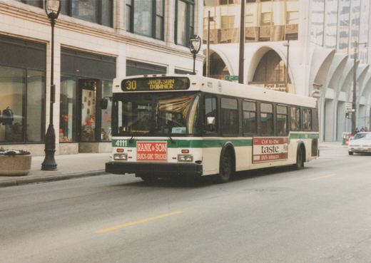 A white and green Flxible Metro bus with the number 4111, operated by Milwaukee County Transit System, is driving on a city street with a '30 Jackson Downer' destination sign.