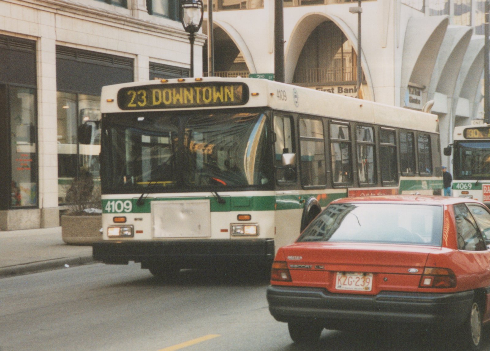 A green and white bus labeled 23 DOWNTOWN and number 4109 is driving on a city street, with a red car in front and another bus behind.