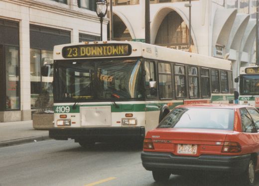 A green and white bus labeled 23 DOWNTOWN and number 4109 is driving on a city street, with a red car in front and another bus behind.