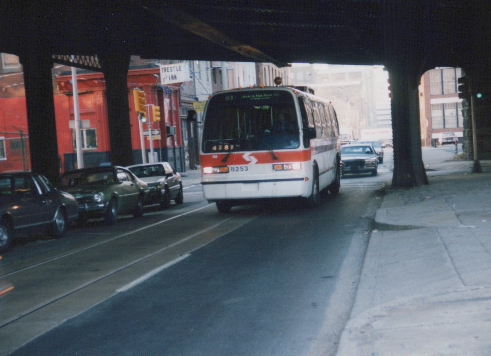 Front view of a white and orange SEPTA bus driving on a city street under a dark elevated structure, with parked cars and a red building in the background.